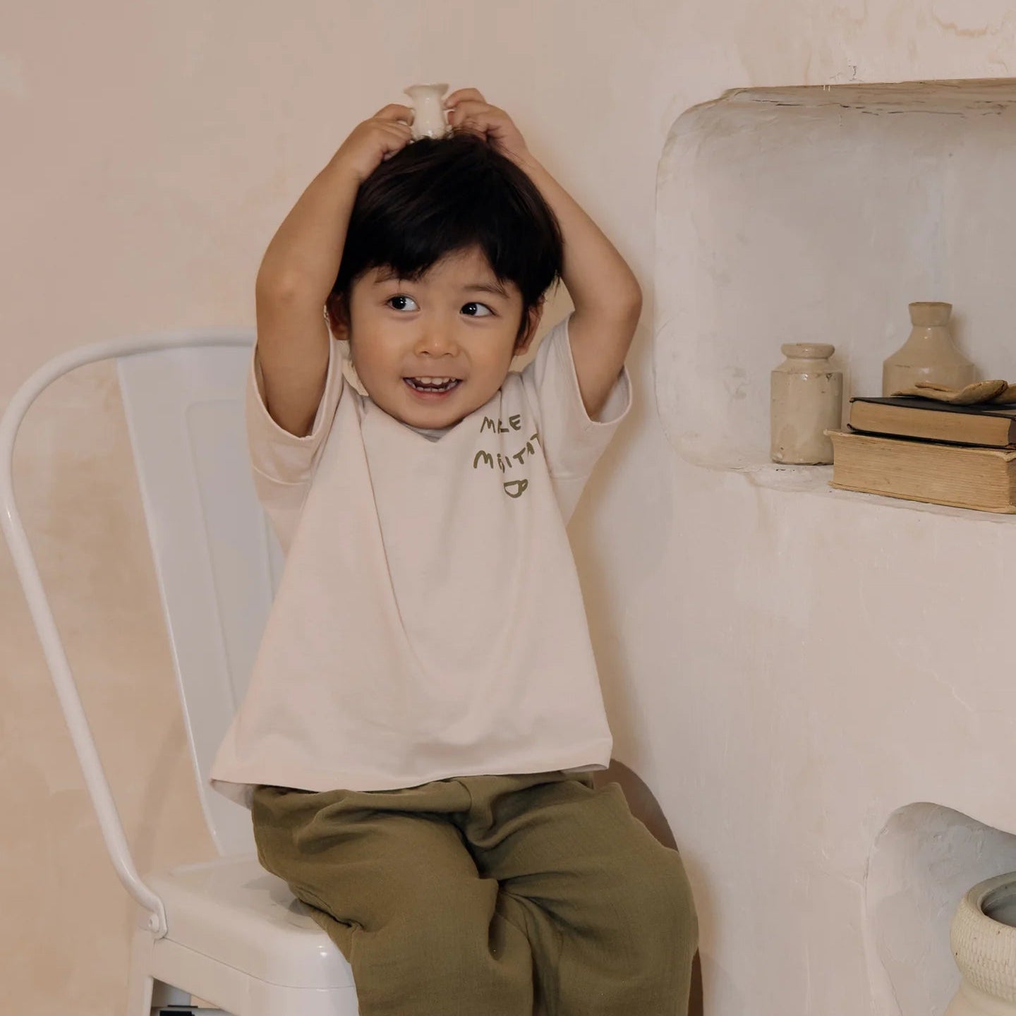 Child sitting on a chair with a white shirt and green pants in a room with a white wall and decorative items.