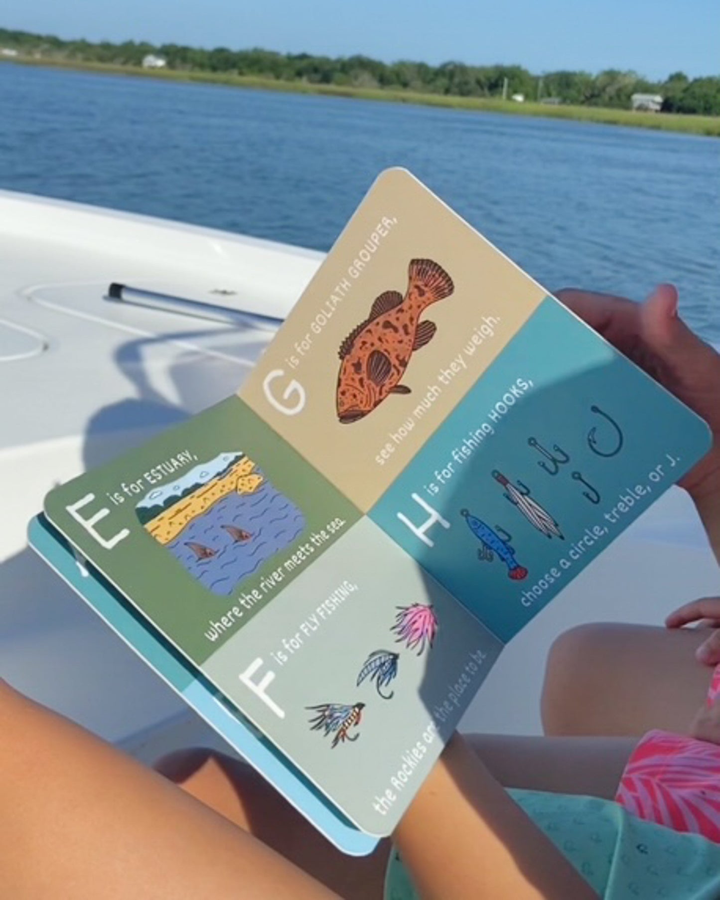 Person holding a colorful book with educational illustrations by a body of water.
