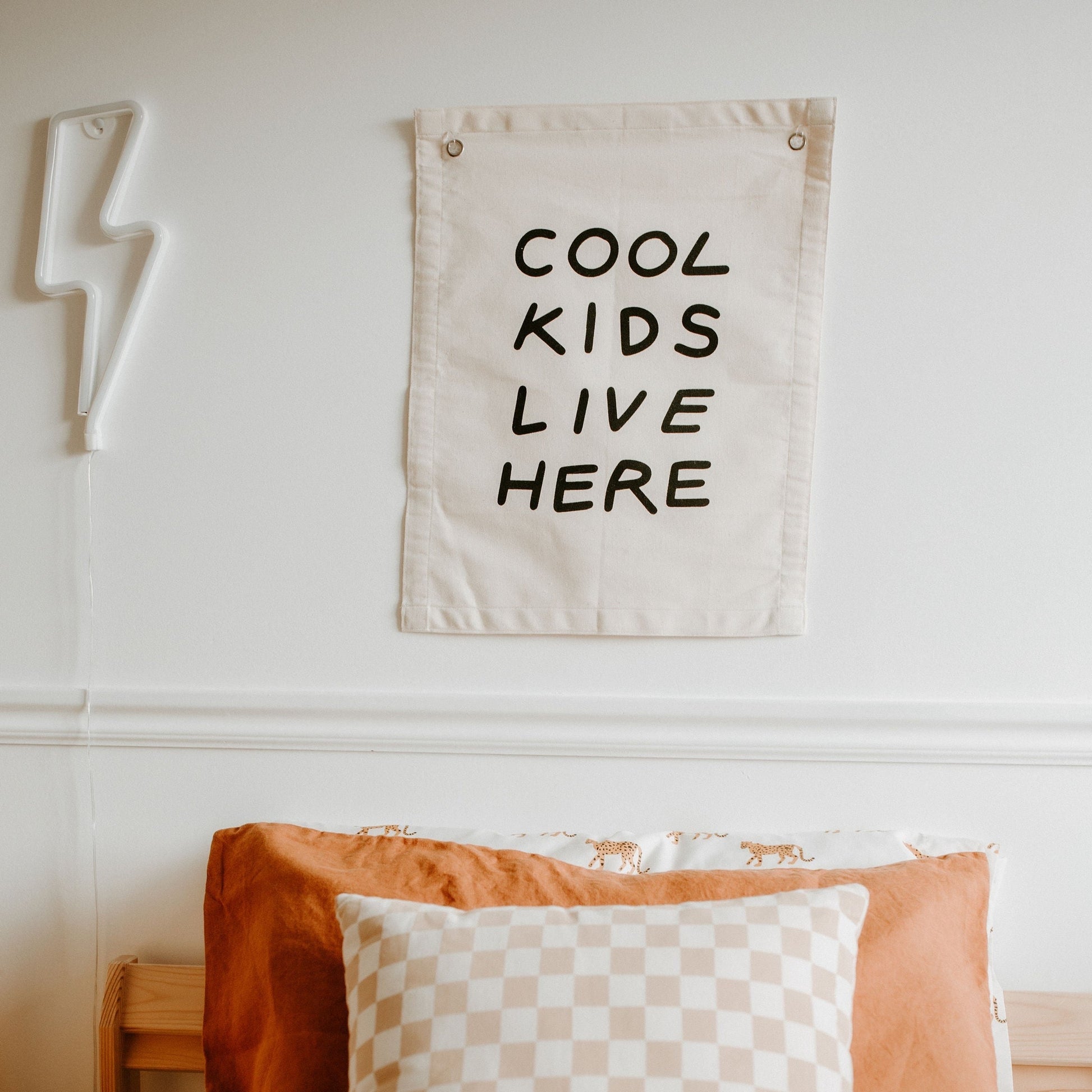 Decorative banner with 'Cool Kids Live Here' text on a wall above a bed with pillows.