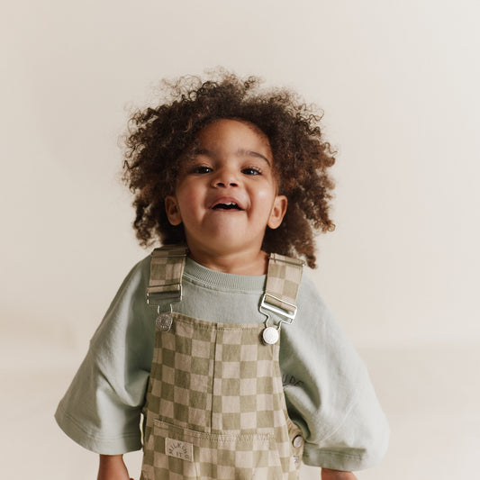Child wearing the Claude & Co. Cecile Olive Checkerboard Dungarees over a mint colored t-shirt with a cream colored backdrop.