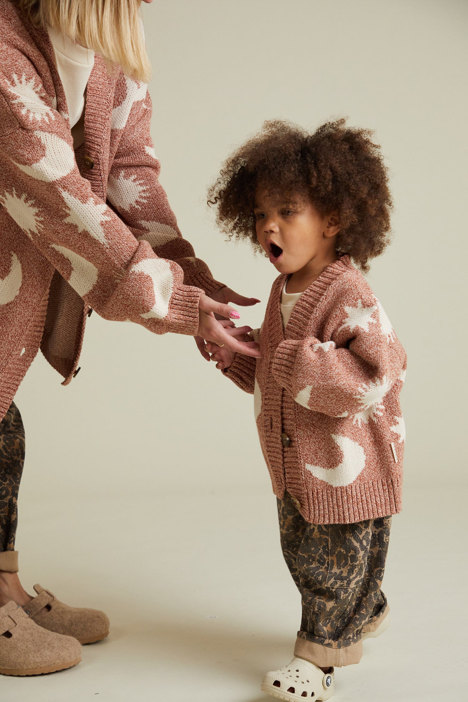 Child wearing a rose colored cardigan with celestial graphics, leopard print trousers, and cream colored crocs. 
