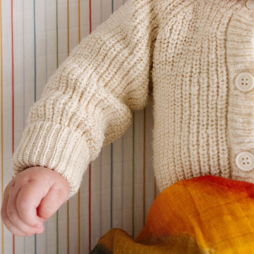 Child wearing a beige knitted sweater with colorful pants against a striped background