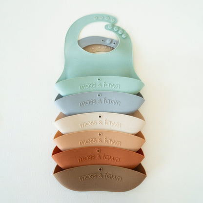 Stack of colorful silicone baby bibs with 'moss & fawn' branding on a light background