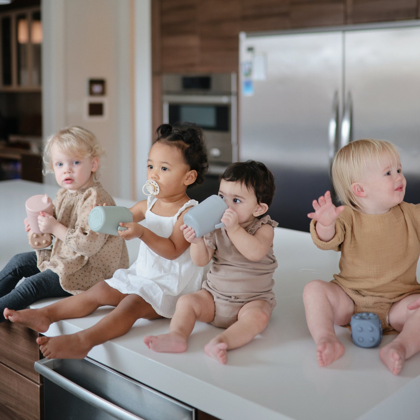 Four toddlers sitting on a kitchen counter with toys, surrounded by kitchen appliances.