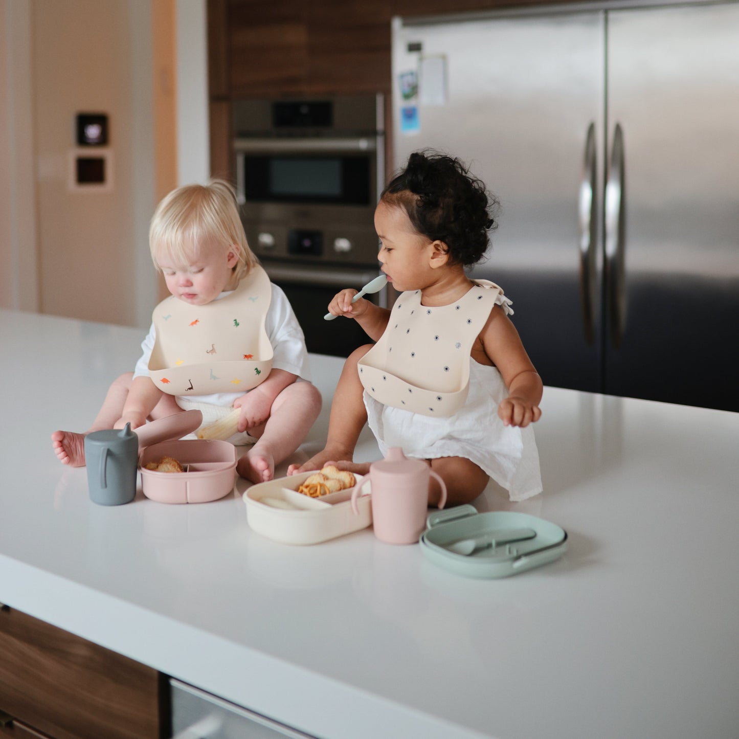 Two children sitting at a kitchen counter with food and utensils, wearing bibs.