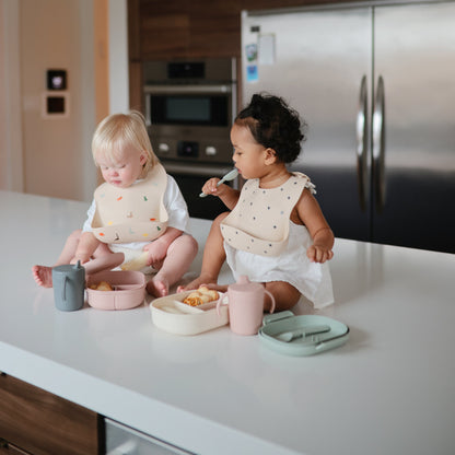Two children sitting at a kitchen counter with food and utensils, wearing bibs.