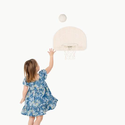 Child in a blue floral dress reaching up to a small basketball hoop on a white background