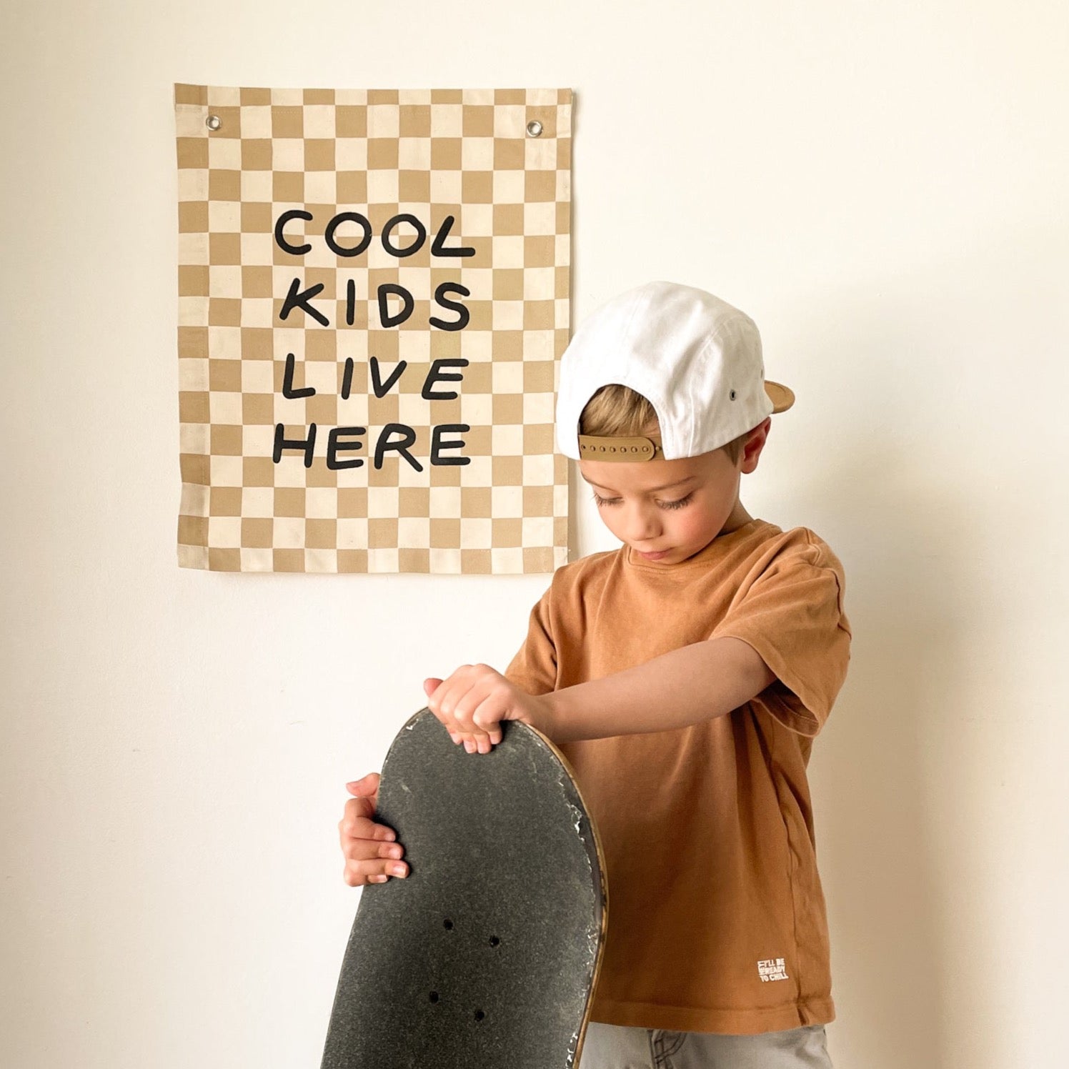 Child holding a skateboard with a 'Cool Kids Live Here' poster on the wall.