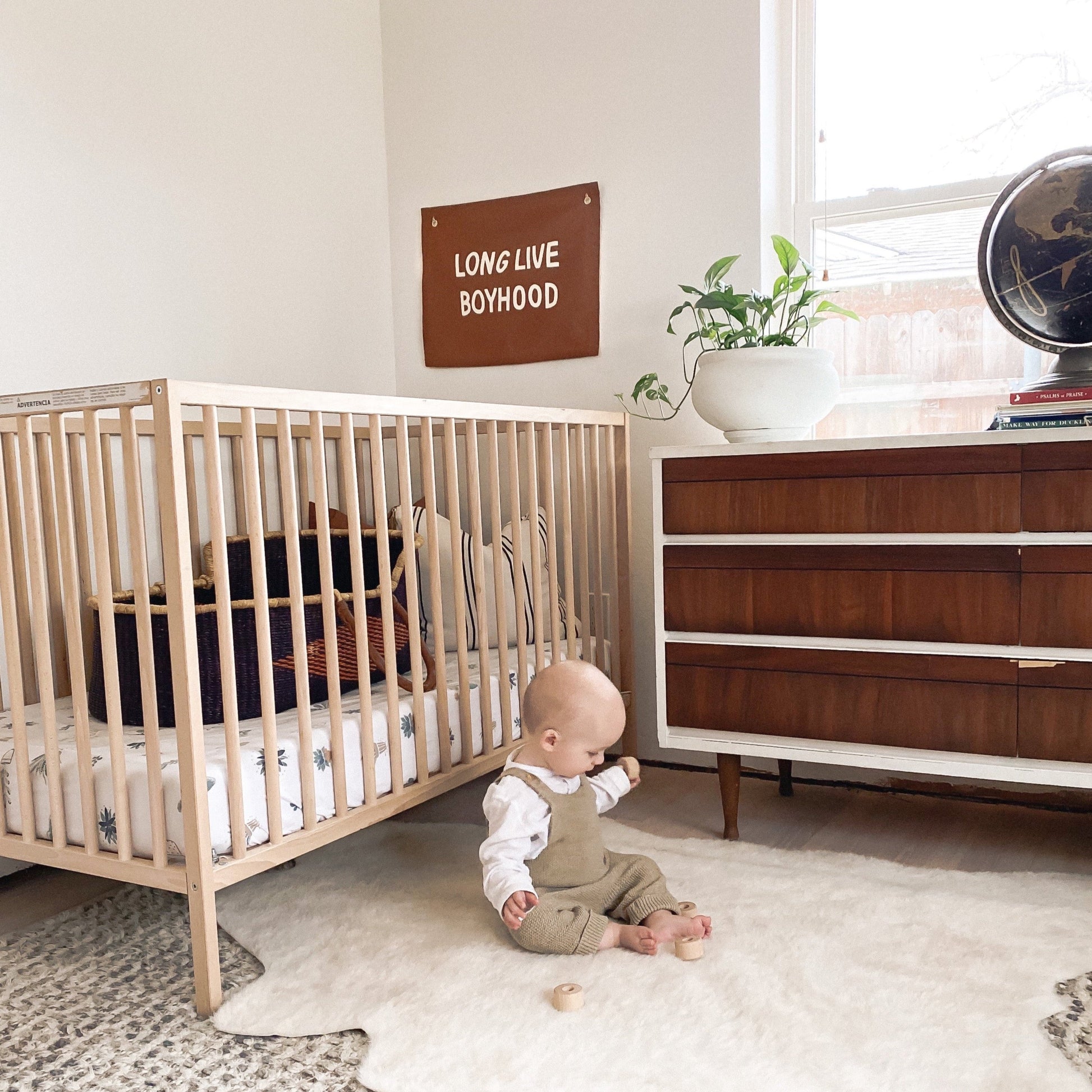 Baby sitting on the floor next to a crib in a room with a dresser and decorative items.
