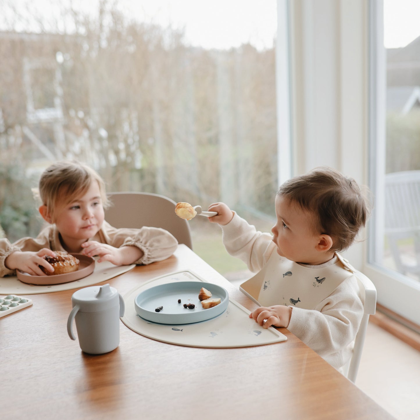 Two children sitting at a table eating snacks in a bright room with large windows.