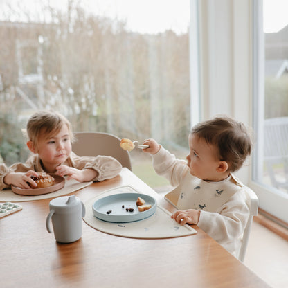 Two children sitting at a table eating snacks in a bright room with large windows.