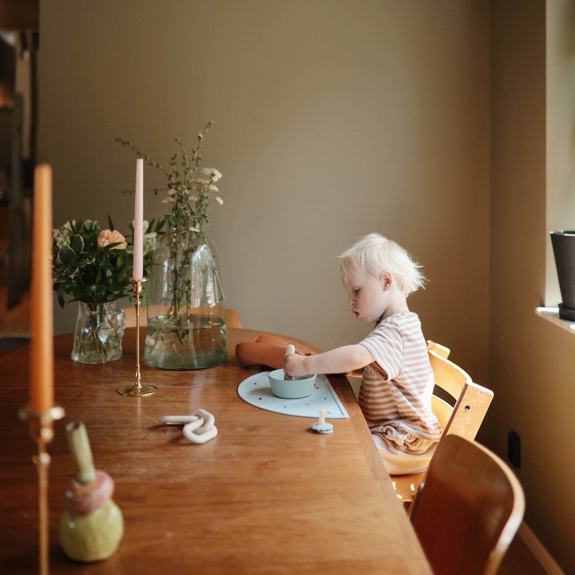 Child sitting at a wooden table with a candle and flowers in a room.