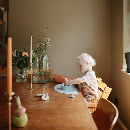 Child sitting at a wooden table with a candle and flowers in a room.