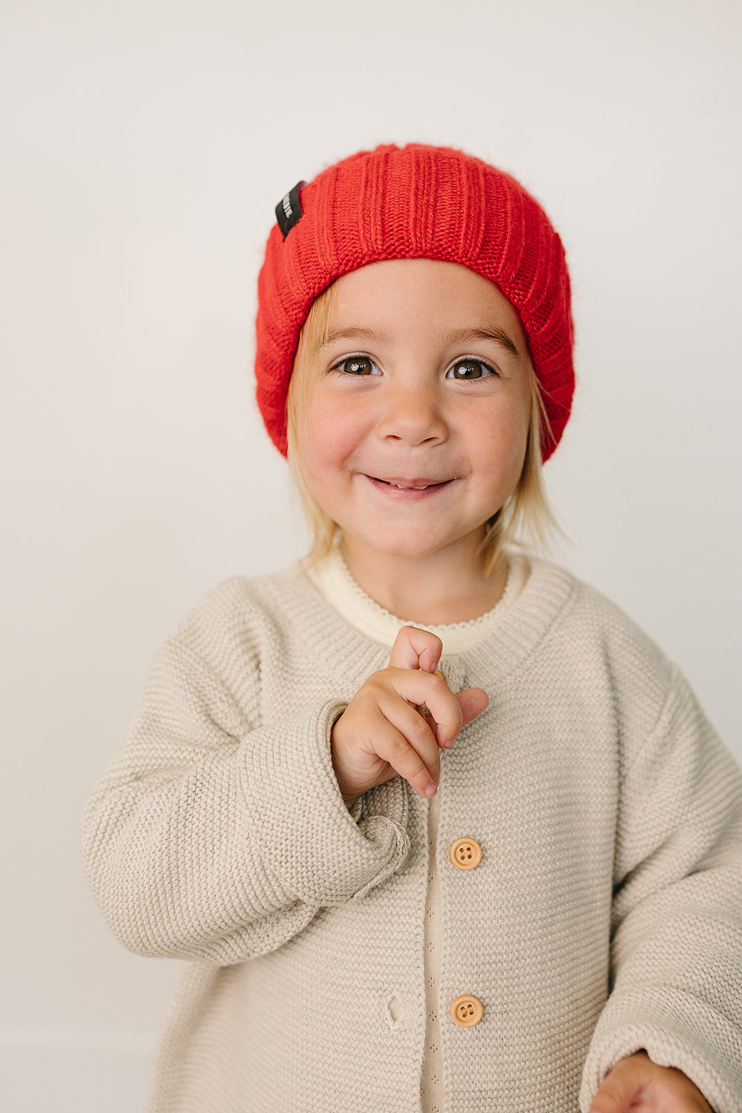 Child wearing a red knit hat and beige cardigan on a plain background