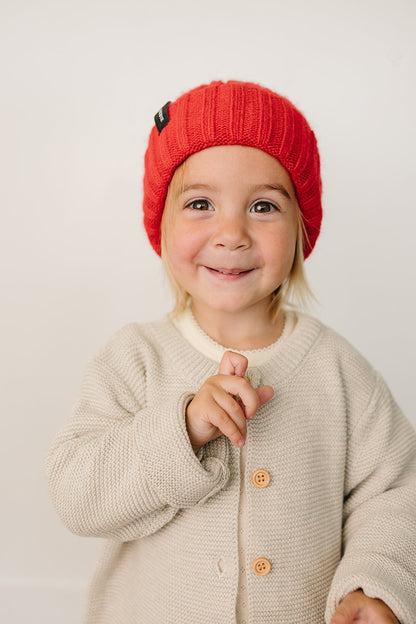 Child wearing a red knit hat and beige cardigan on a plain background