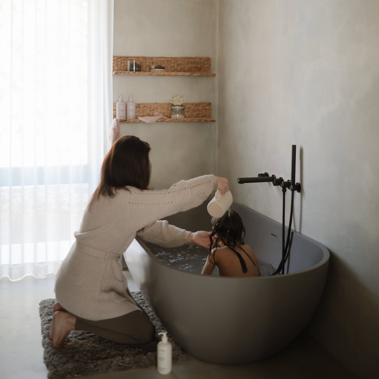 Woman in a beige robe helping another person in a bathtub with a white bottle.