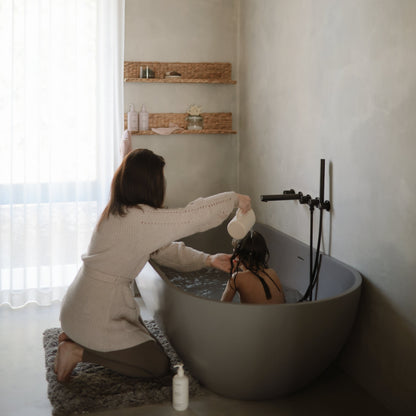 Woman in a beige robe helping another person in a bathtub with a white bottle.