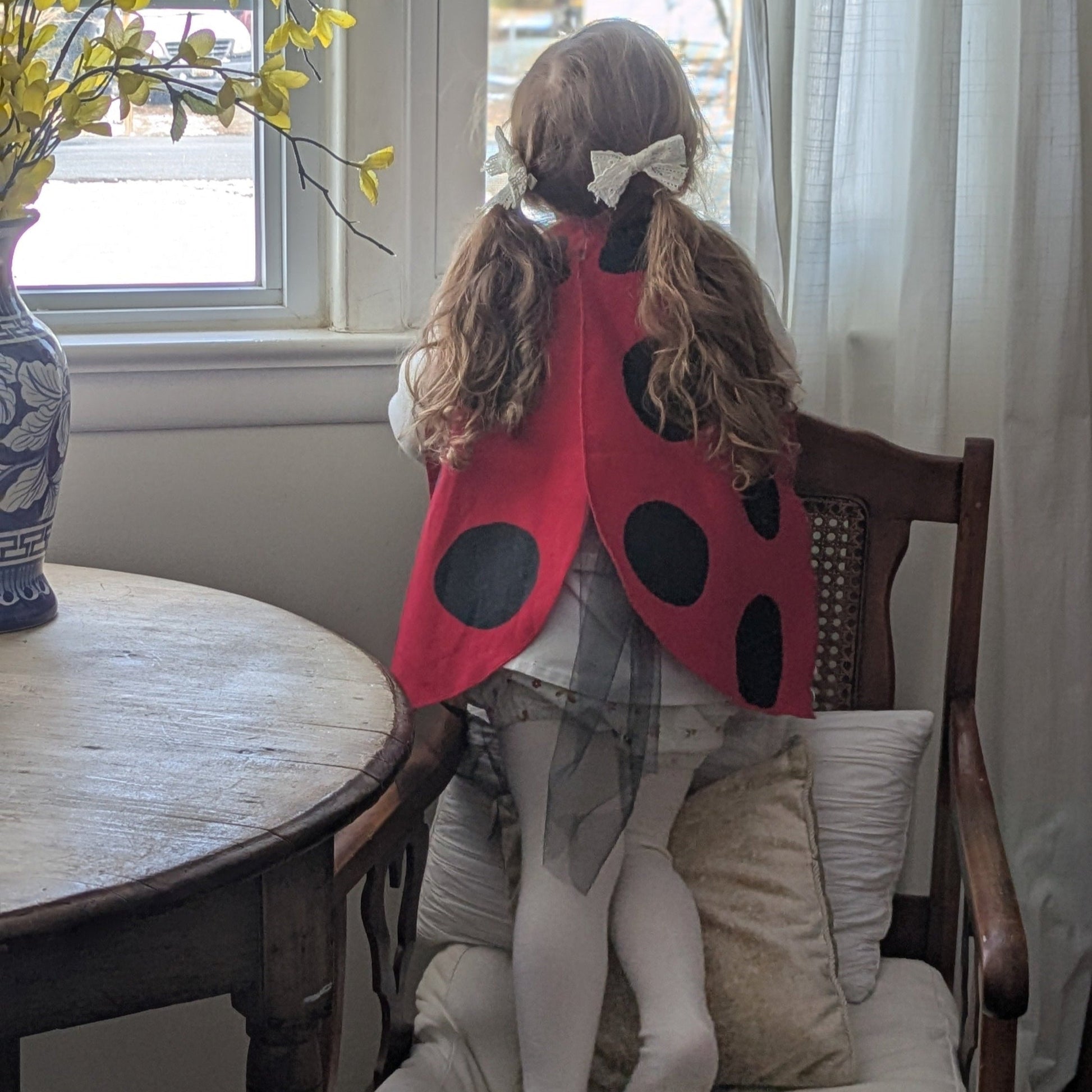Child wearing a ladybug costume sitting on a chair in a room with a table and window.