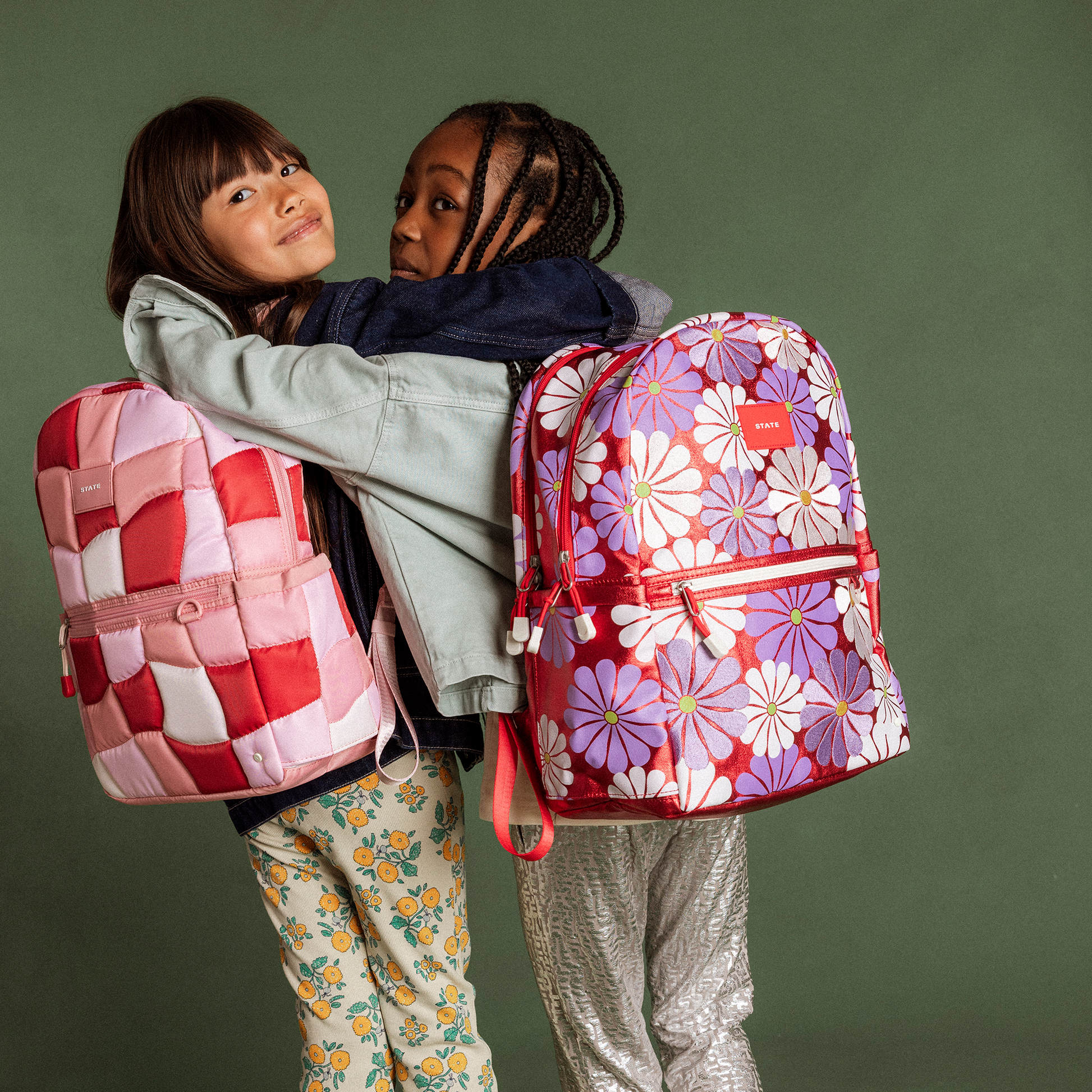 Two children hugging with colorful backpacks against a green background
