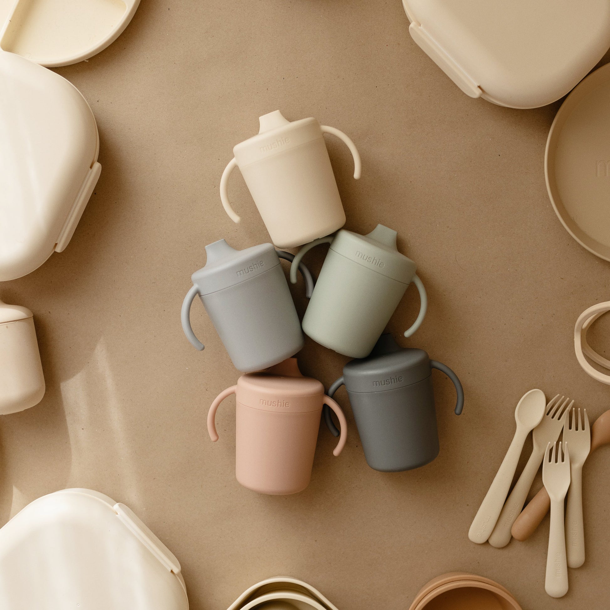 Collection of colorful children's cups, bowls, and utensils on a beige surface