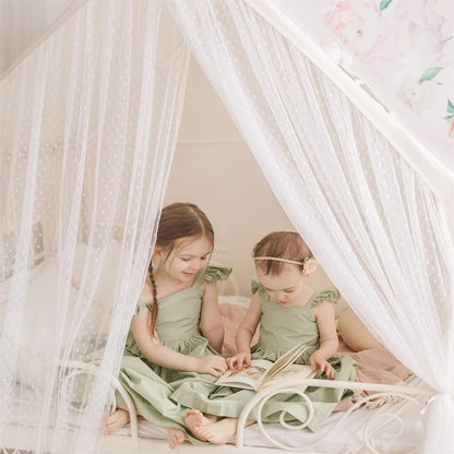 Two young girls in green dresses sitting inside a white canopy bed, reading a book.