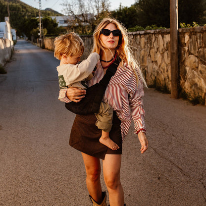 Woman holding a child in a sling on a street with stone walls and trees in the background
