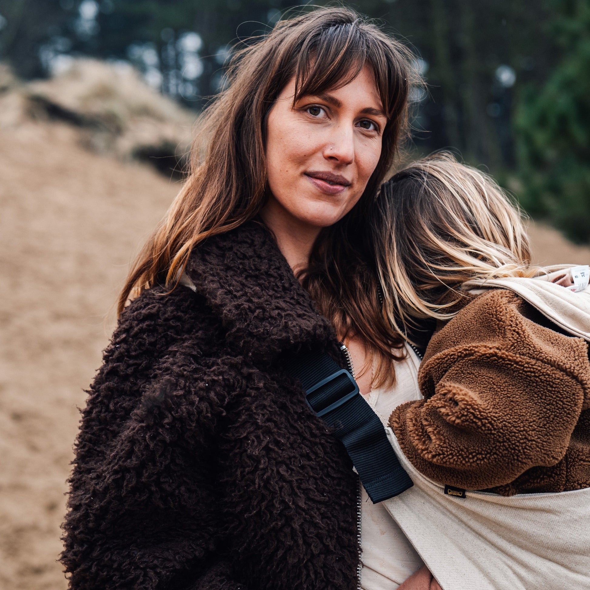 Woman holding a child in a sling outdoors with trees in the background