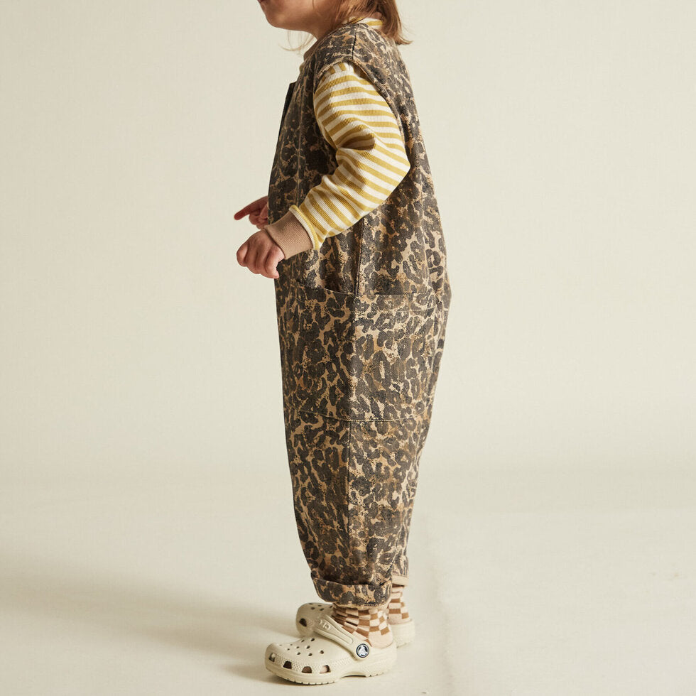 A child standing side-on wearing a sleeveless, leopard print overall with tortoiseshell style buttons and oversized pockets.