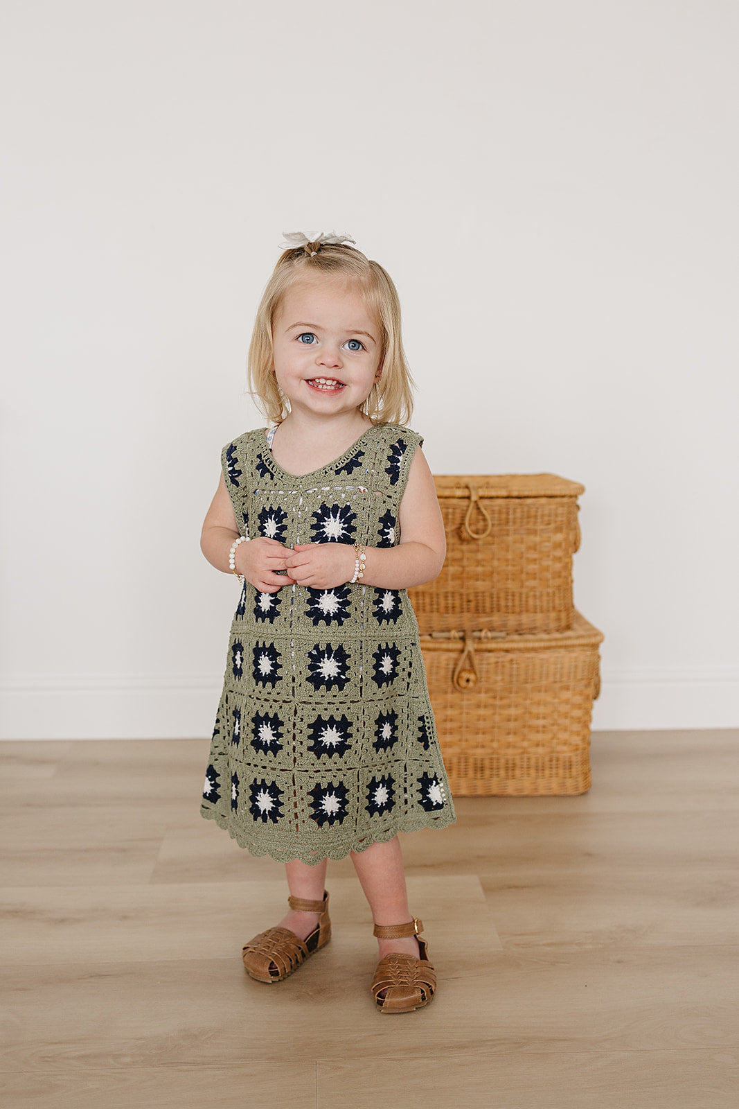 Young girl wearing a green crochet dress with a floral pattern, standing in front of wicker baskets.