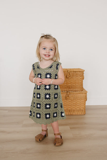 Young girl wearing a green crochet dress with a floral pattern, standing in front of wicker baskets.