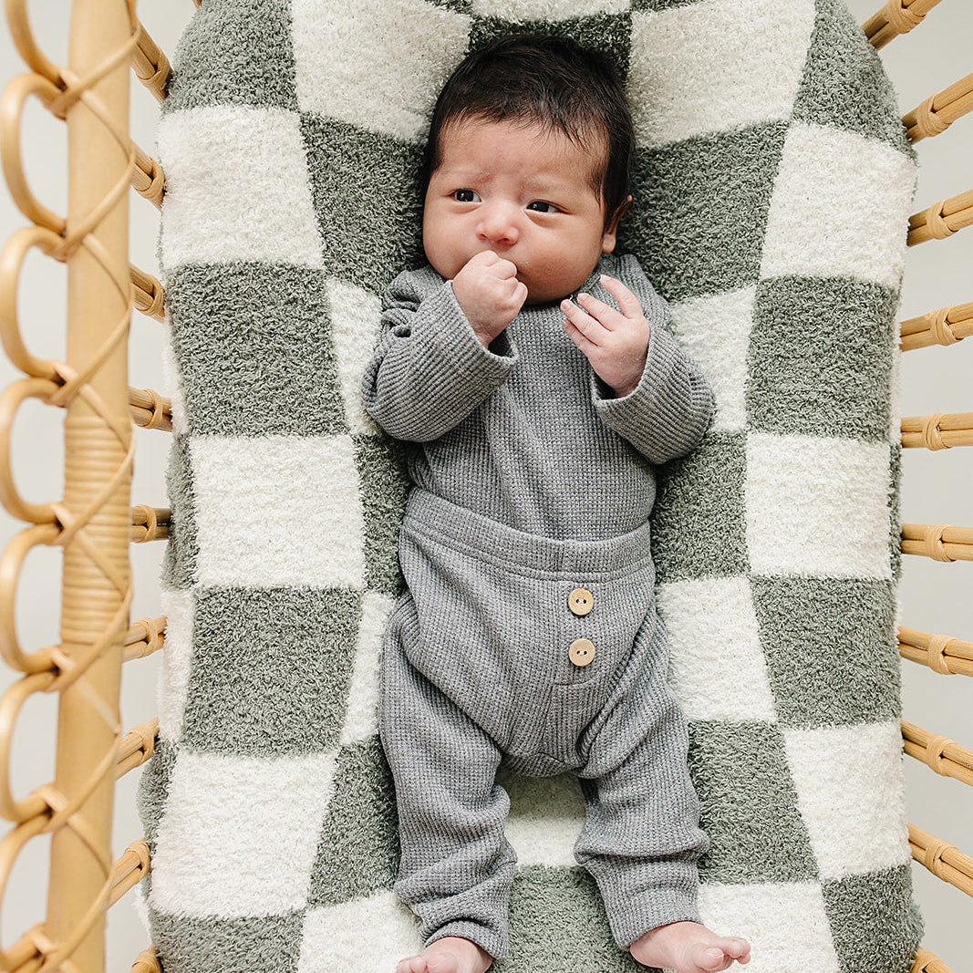 Newborn baby in a gray outfit lying on a checkered blanket in a wicker basket.