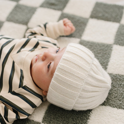 Baby wearing a white knit hat and striped outfit lying on a checkered rug.