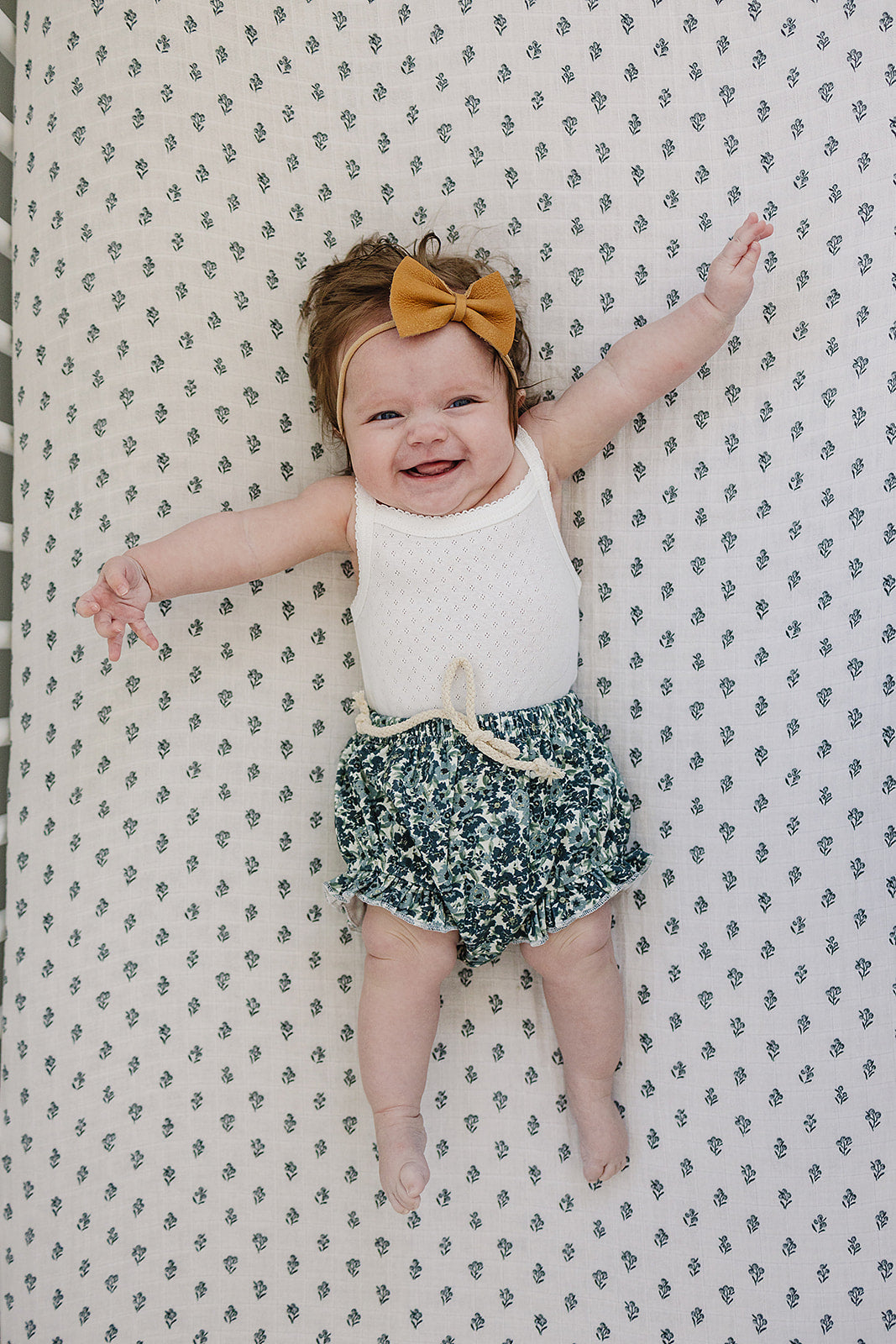 Baby lying on a patterned surface wearing a white top and green floral shorts with a yellow bow.