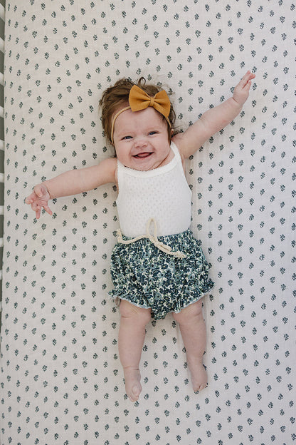 Baby lying on a patterned surface wearing a white top and green floral shorts with a yellow bow.