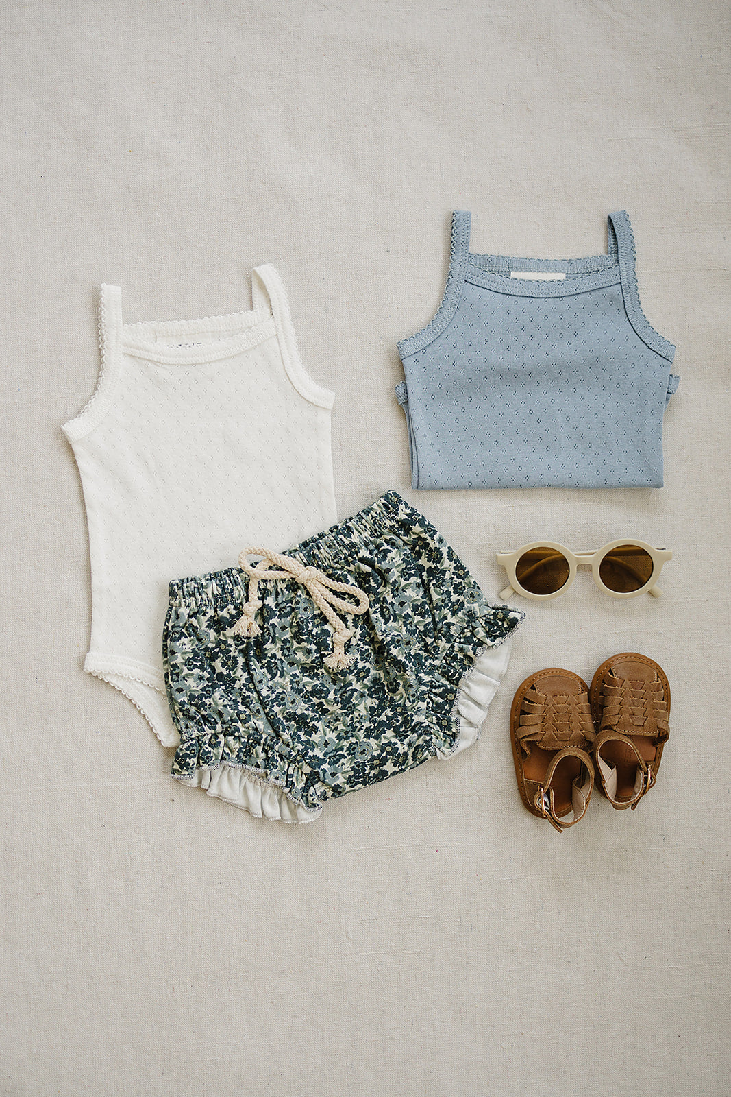 Children's clothing set including a white tank top, floral shorts, blue tank top, brown sandals, and sunglasses on a beige background.