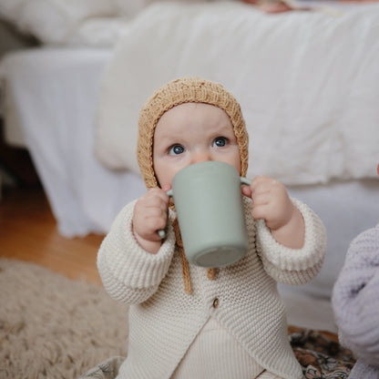 Baby in a knitted outfit and hat holding a green mug indoors.