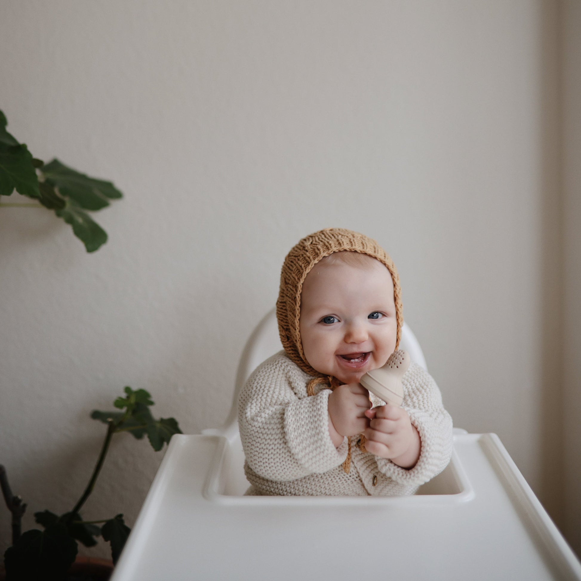 Baby in a knit outfit and bonnet sitting in a high chair with a plant in the background