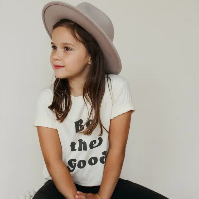 Young girl sitting on a rug wearing a white t-shirt with text and a gray hat.