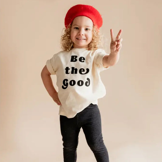 Child wearing a white t-shirt with 'Be the Good' text, red beret, and black pants on a beige background