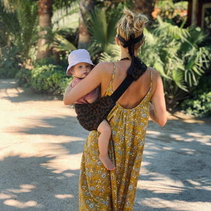 Woman in a yellow floral dress carrying a baby in a leopard print sling through a tropical setting.