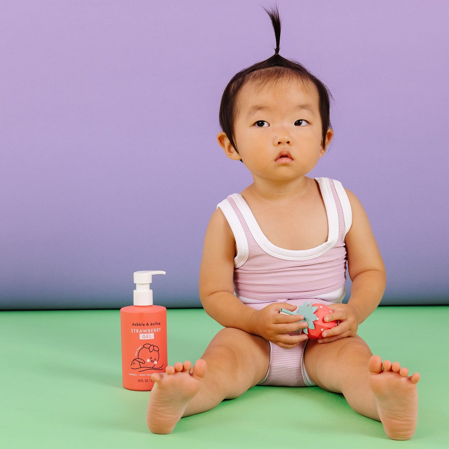 Child sitting on a green floor with a pink bottle and toy, against a purple background