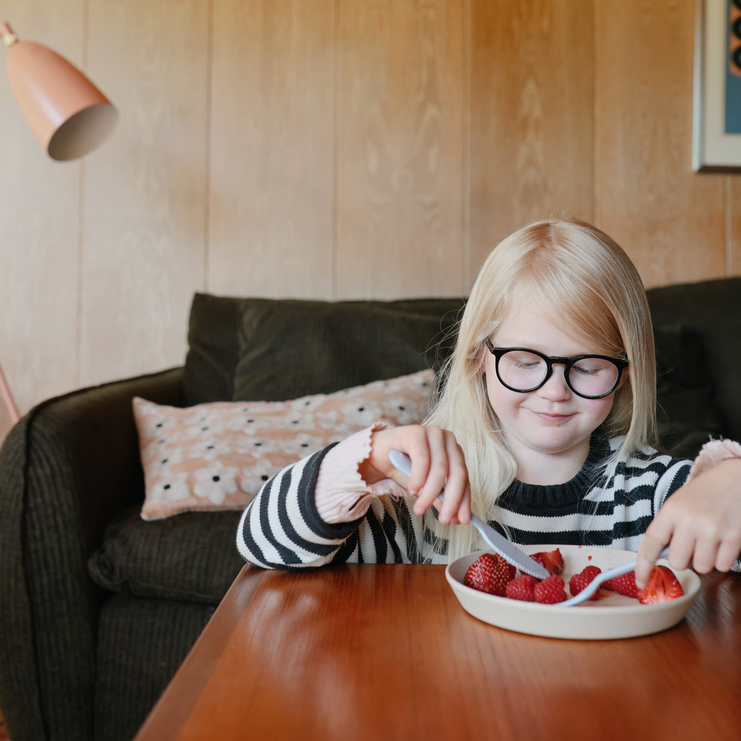 Child with glasses eating strawberries at a table in a cozy living room.