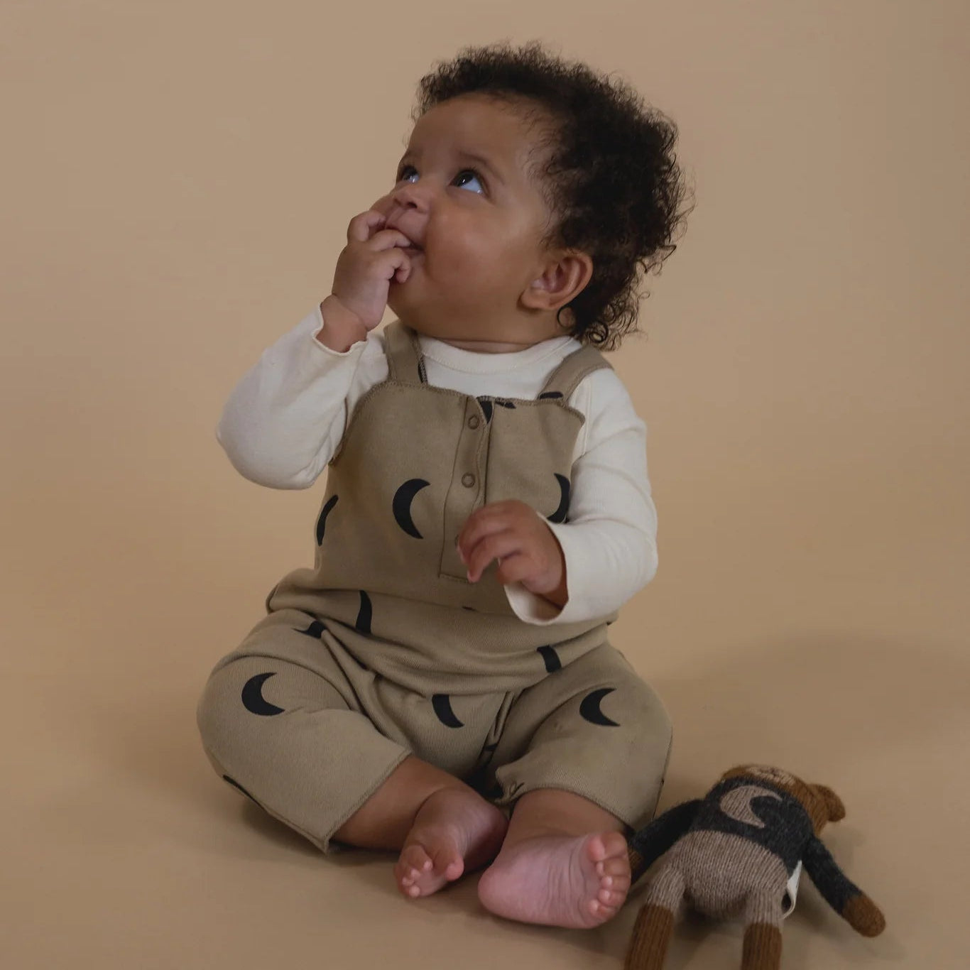 Baby in beige romper with black moon patterns, sitting on a beige background with a plush toy.