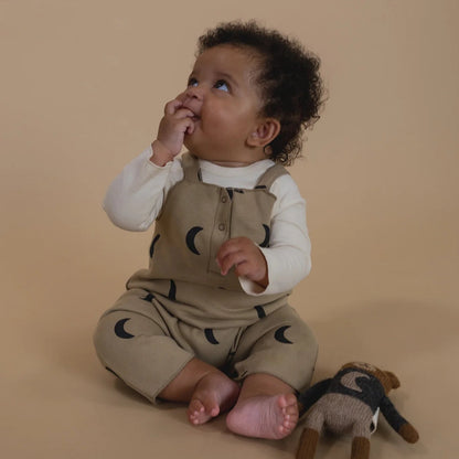 Baby in beige romper with black moon patterns, sitting on a beige background with a plush toy.