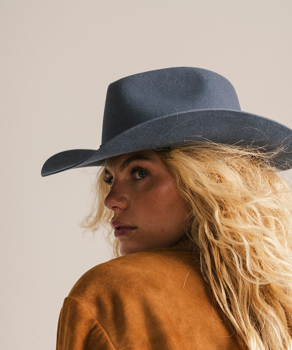 Person wearing a blue cowboy hat and brown jacket against a plain background