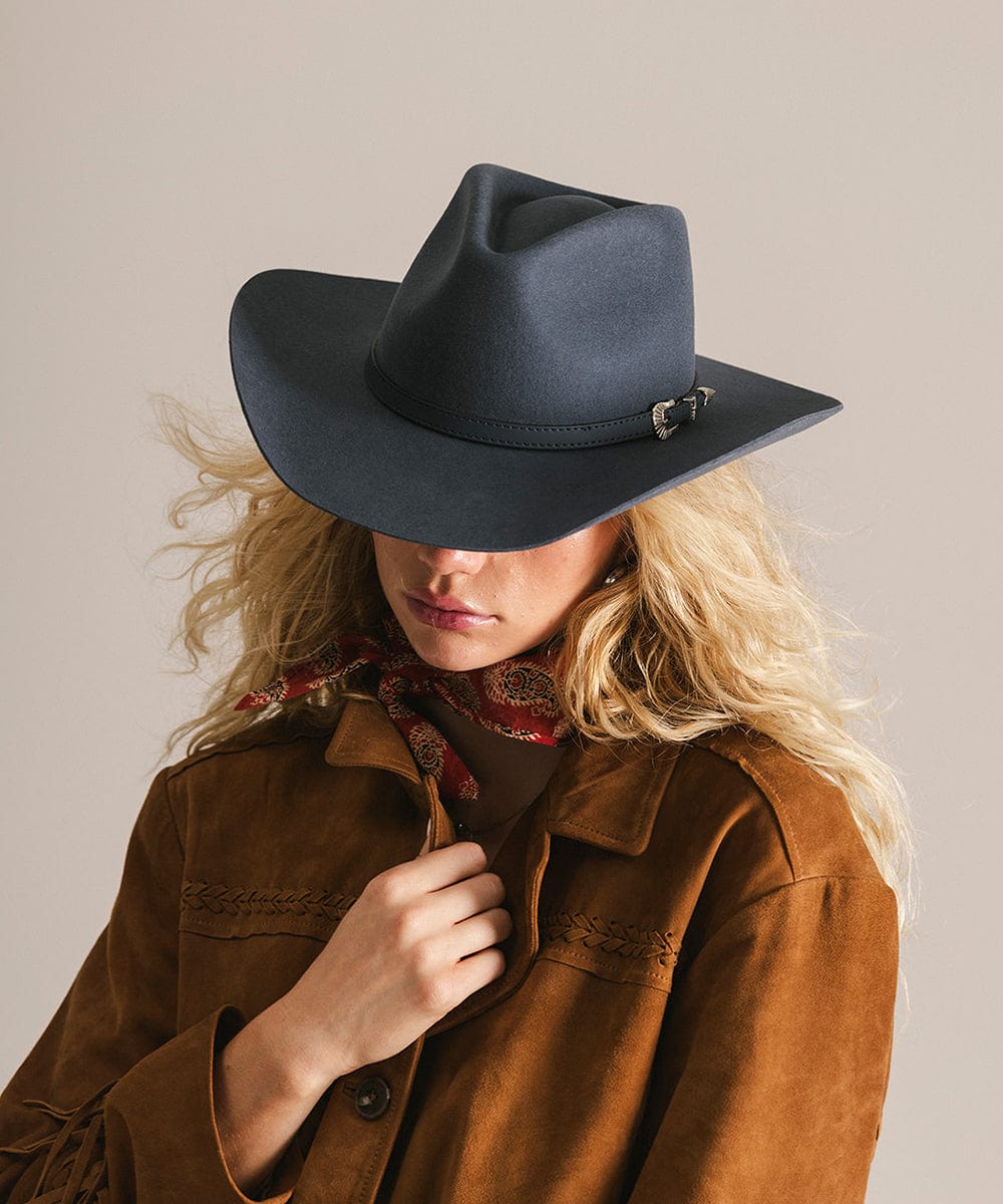 Person wearing a dark blue cowboy hat and brown jacket with a patterned scarf on a neutral background