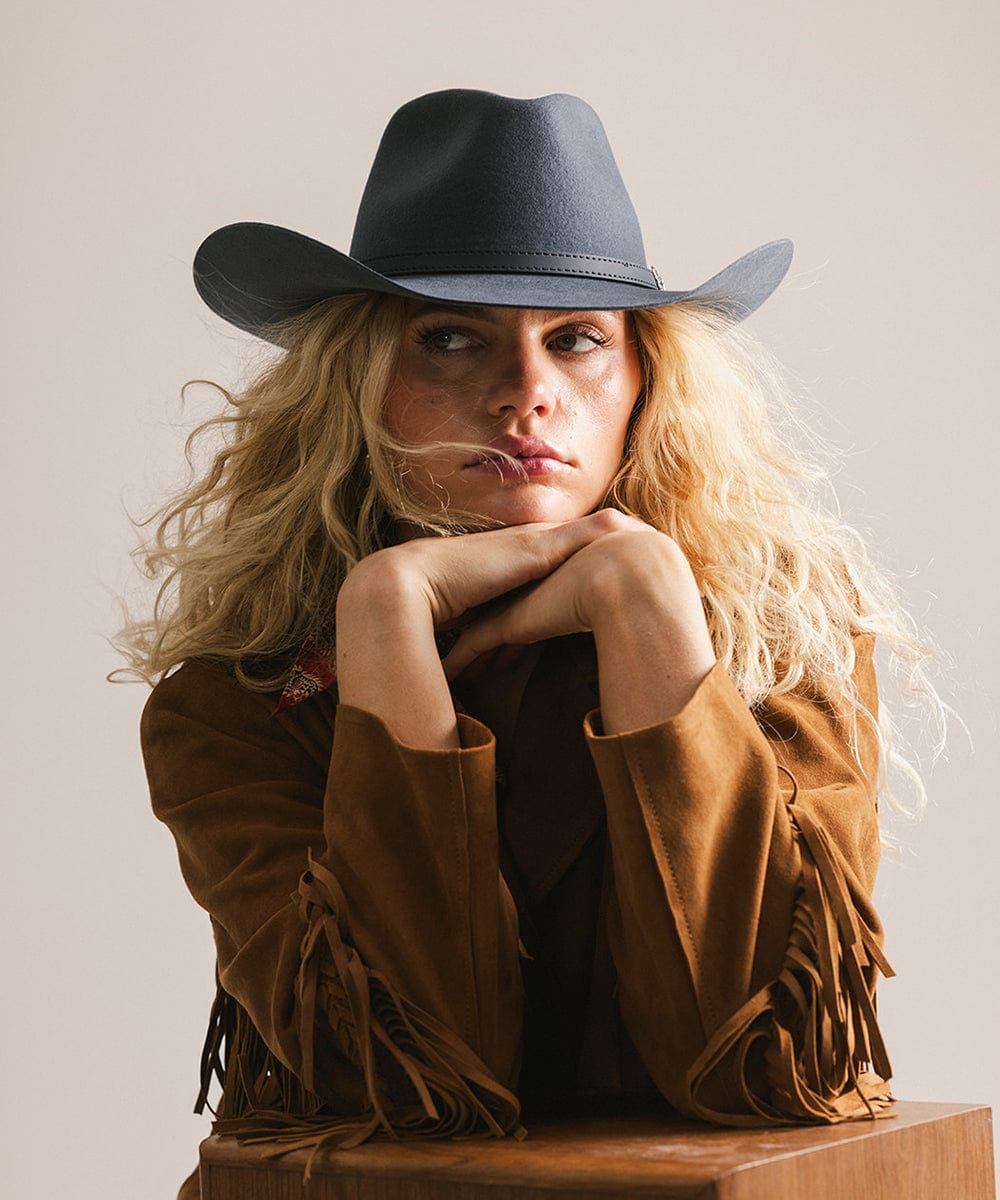 Woman wearing a cowboy hat and brown jacket with fringe, sitting on a wooden block against a plain background