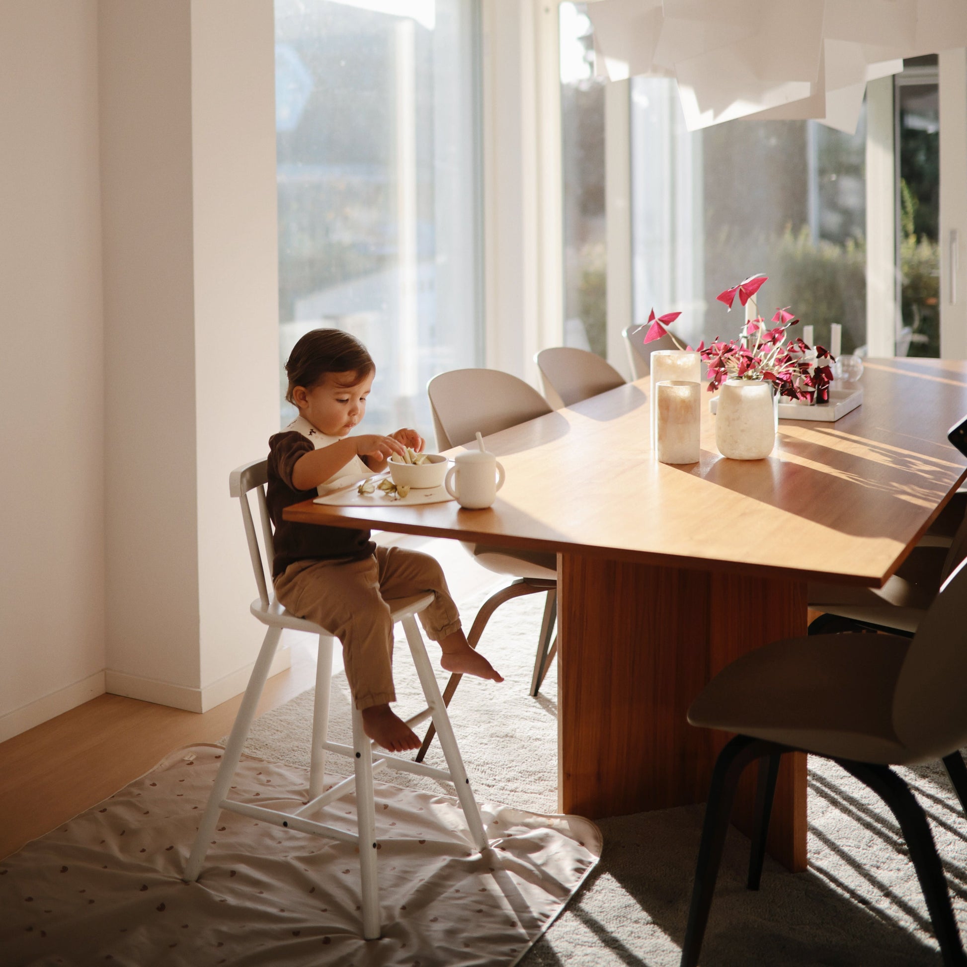 Child sitting at a wooden dining table in a bright room with large windows.