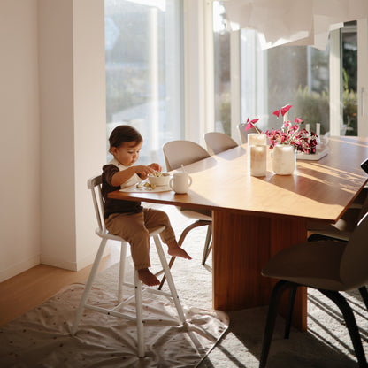 Child sitting at a wooden dining table in a bright room with large windows.