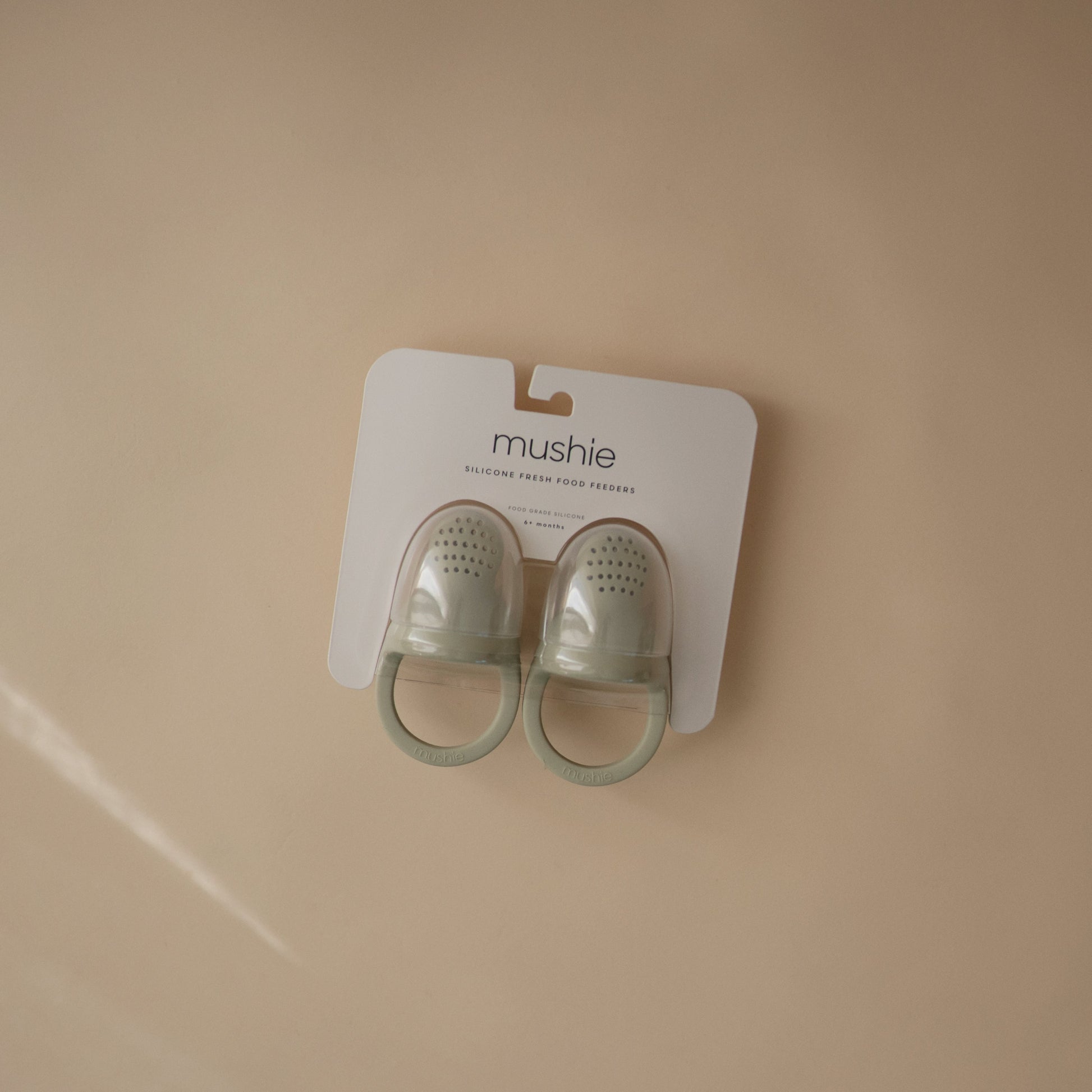 Pair of green baby shoes on a Mushie branded card against a beige background